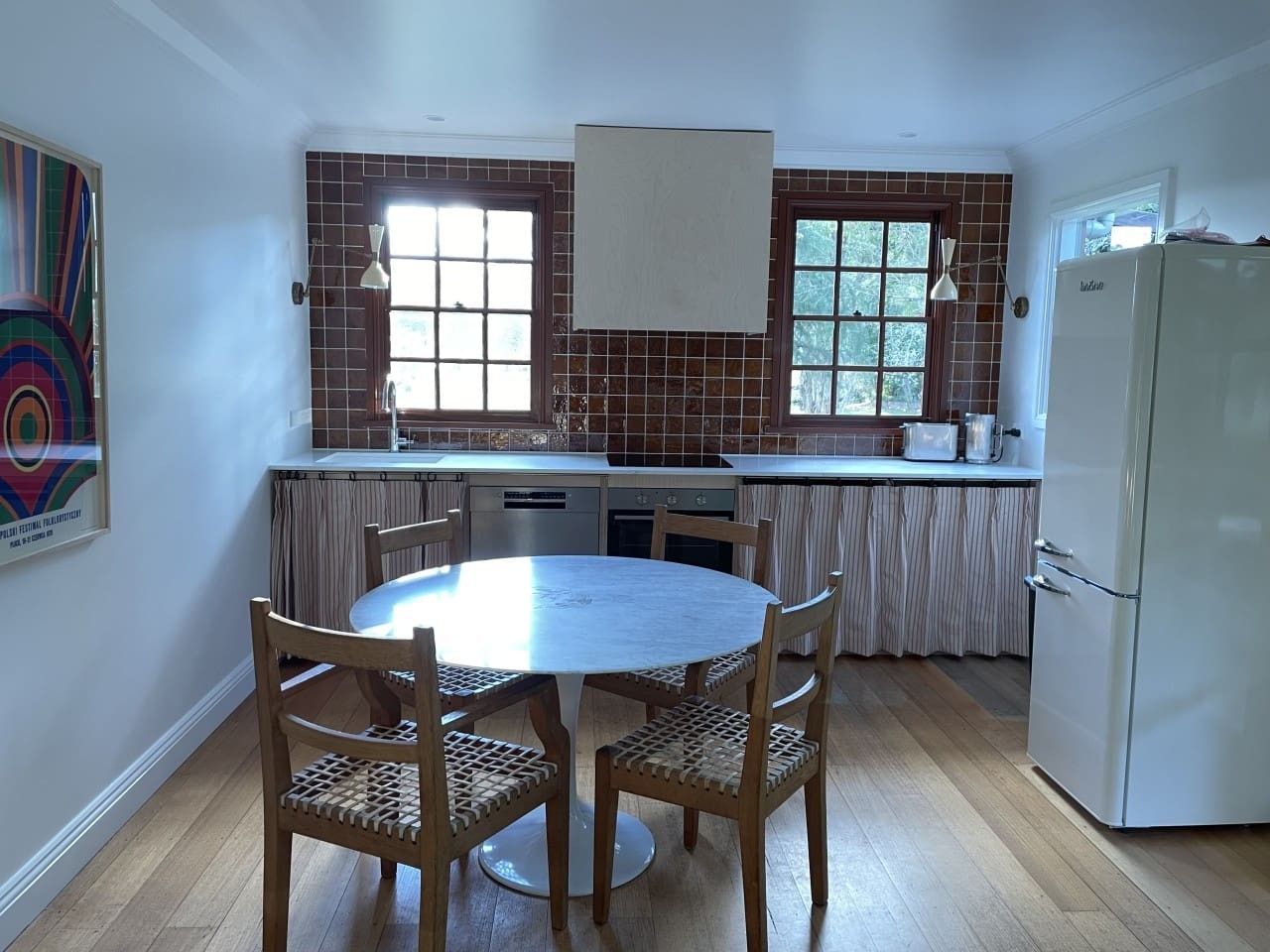A kitchen with a round dining table, wooden chairs, a refrigerator, and a window with curtains.