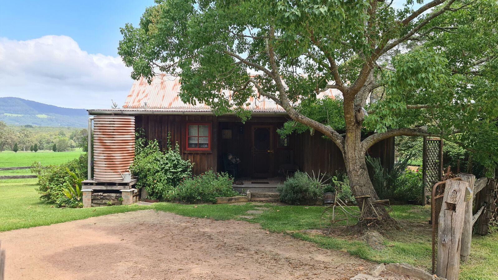 A rustic wooden house with a metal roof, surrounded by greenery and a dirt path leading to the entrance.