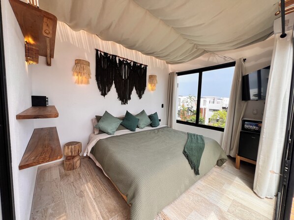 A modern bedroom with a large bed, wooden shelves, a wall-mounted TV, and a view of buildings through a large window. - New 3BR PH Jacuzzi, Views, GYM, Pool, Cinema, WiFi (Tulum)