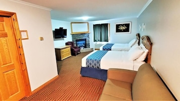 Habitación de hotel con puerta de madera, televisor, chimenea, cama con colcha azul y blanca, sofá y escritorio pequeño con una silla.