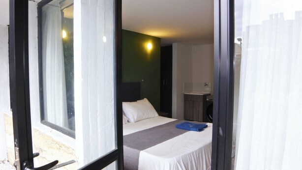 A hotel room with a bed, a small table, a bathroom with a sink, and a view through a glass door. - ApartaHotel Retiro 79 (Bogotá)