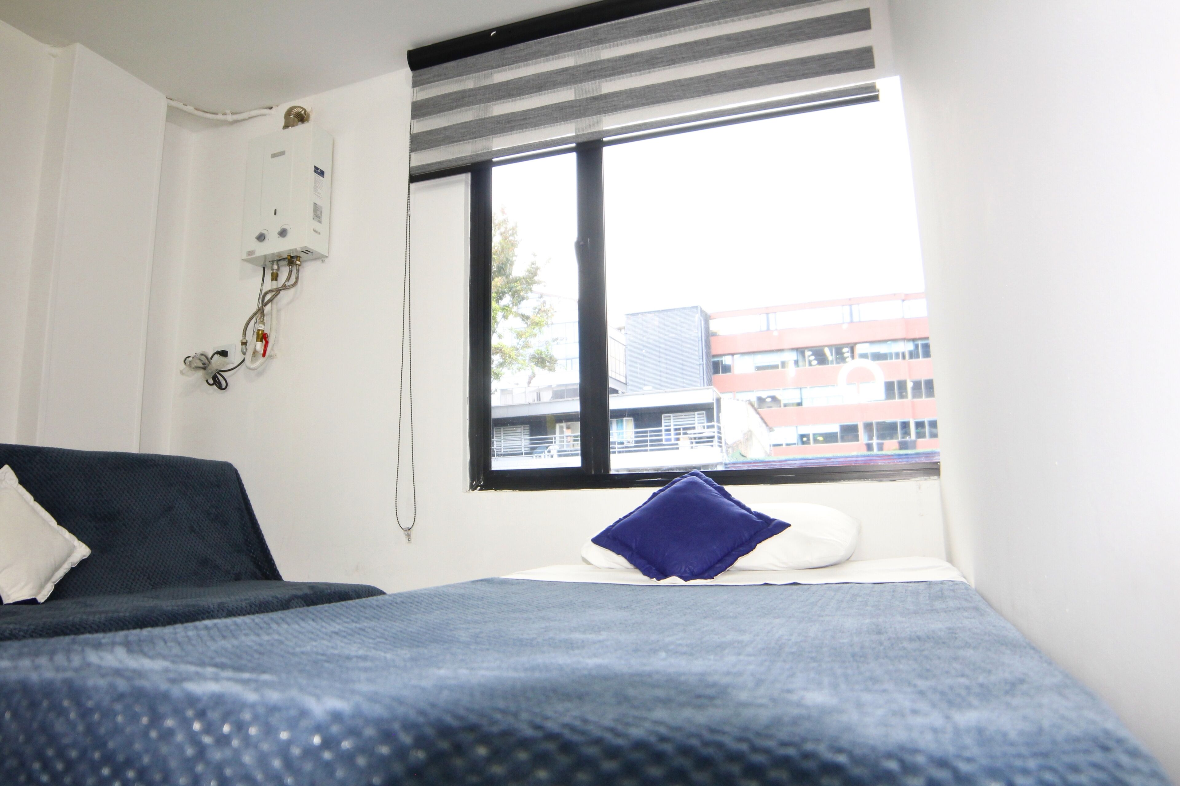 Habitación de hotel con cama, ventana con persianas, termostato en la pared y vista a edificios.