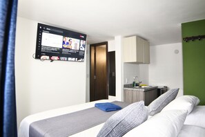Quarto de hotel com cama, televisão presa à parede, pequeno espaço de cozinha e banheiro visível através de uma porta aberta.