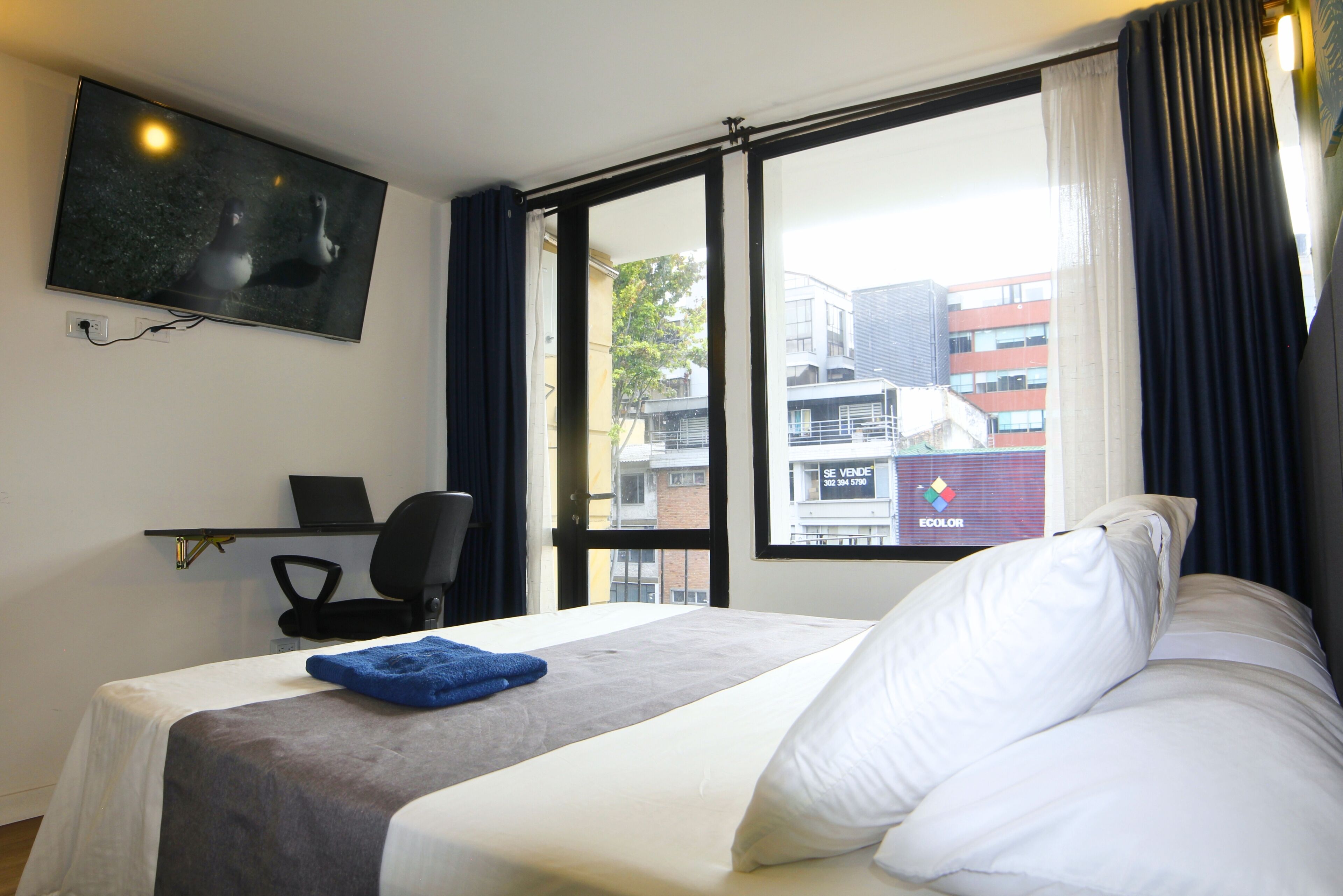 A hotel room with a bed, a desk with a chair, a TV mounted on the wall, and a window with curtains.