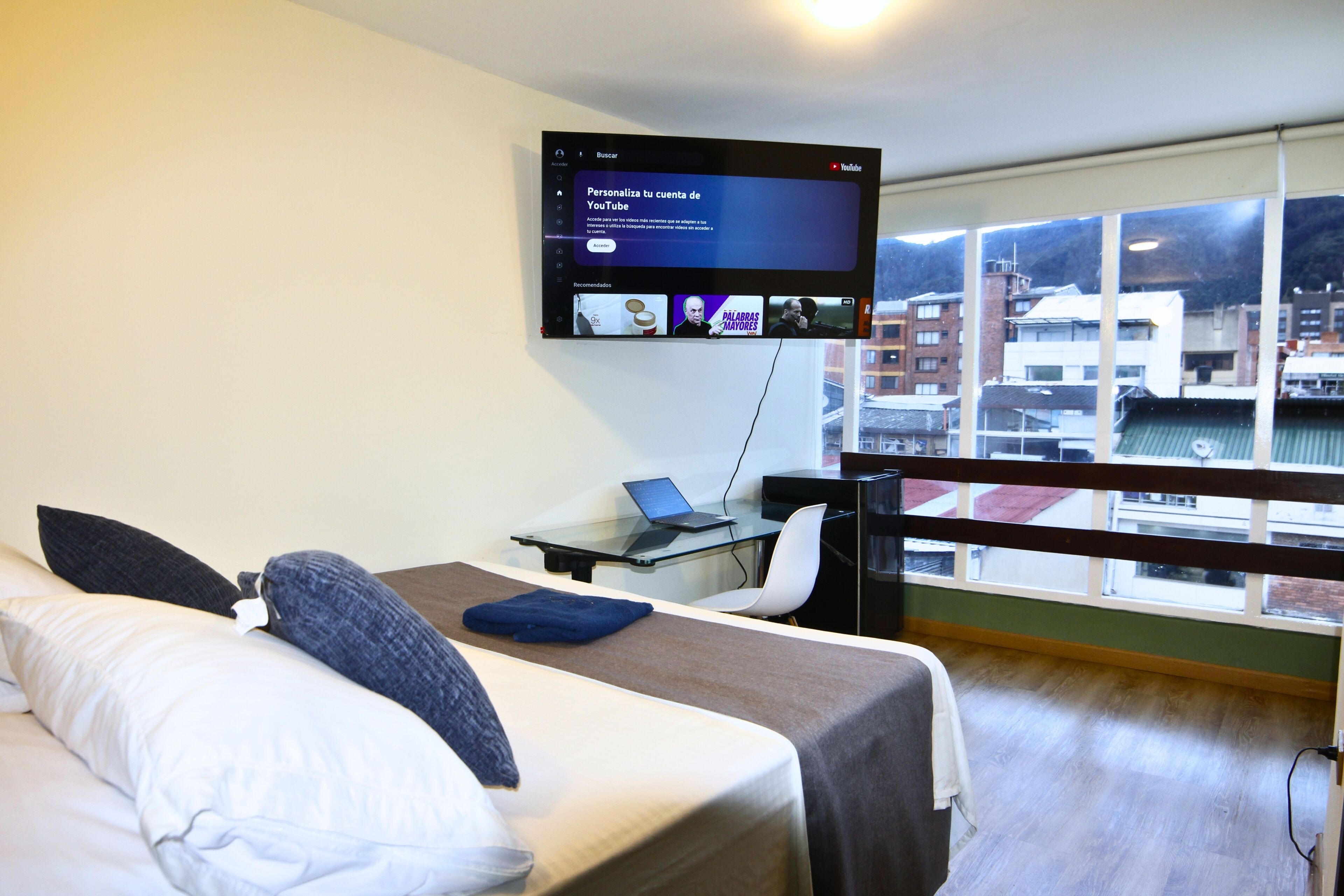 Une chambre d’hôtel avec un lit, un bureau avec un ordinateur portable, un téléviseur fixé au mur et une vue sur les immeubles à travers de grandes fenêtres.