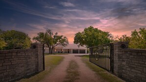 A gated driveway leading to a building with a garage, surrounded by trees and a clear sky. - Rivers Oaks-the Ranch House Pool & Hot-tub! (Fredericksburg)