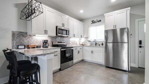 A modern kitchen with white cabinets, stainless steel appliances, a herringbone tile backsplash, and a bar area with stools. - Pet Friendly W/hot-tub-six Minutes to Downtown! (Fredericksburg)