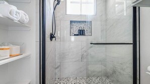 A modern bathroom with a glass-enclosed shower, marble tiles, a towel rack, and a shelf with toiletries. - Pet Friendly W/hot-tub-six Minutes to Downtown! (Fredericksburg)