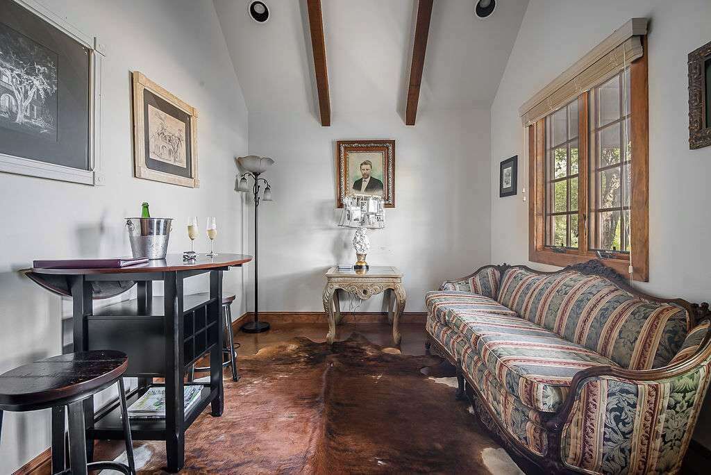 Una habitación con piso de madera, un sofá estampado, una mesa con una lámpara y cuadros en las paredes.