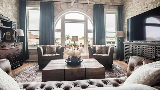 A spacious living room with a large arched window, leather sofas, a coffee table with a vase, a television, and a stone wall. - The Bank-luxury Suite W/deck Overlooking Main St! (Fredericksburg)