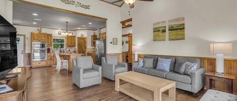 A spacious living area with a grey sofa, two armchairs, a wooden coffee table, and a ceiling fan. A kitchen with wooden cabinets and stainless steel appliances is visible in the background.
