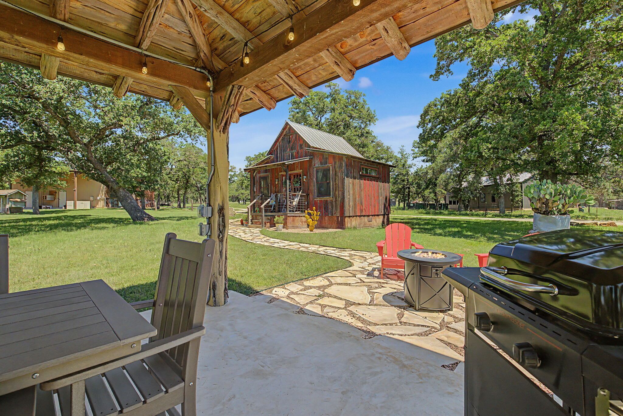 Een terras buiten met een houten pergola, een barbecue en een pad van stenen dat naar een rustiek gebouw leidt.