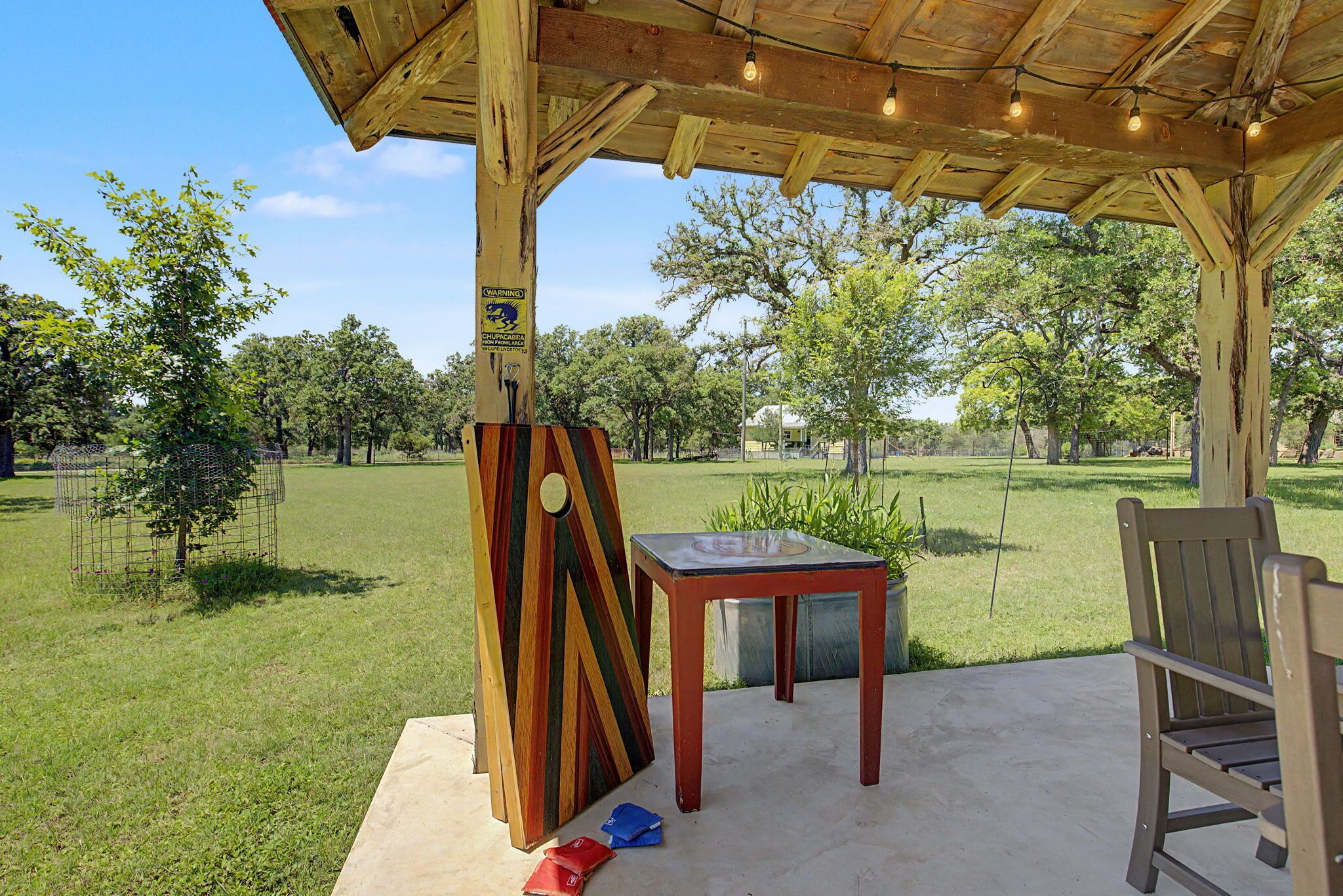 Un espace extérieur aménagé avec une table et des chaises en bois, un panneau d’avertissement et une zone délimitée par une clôture, où se trouvent des arbres.