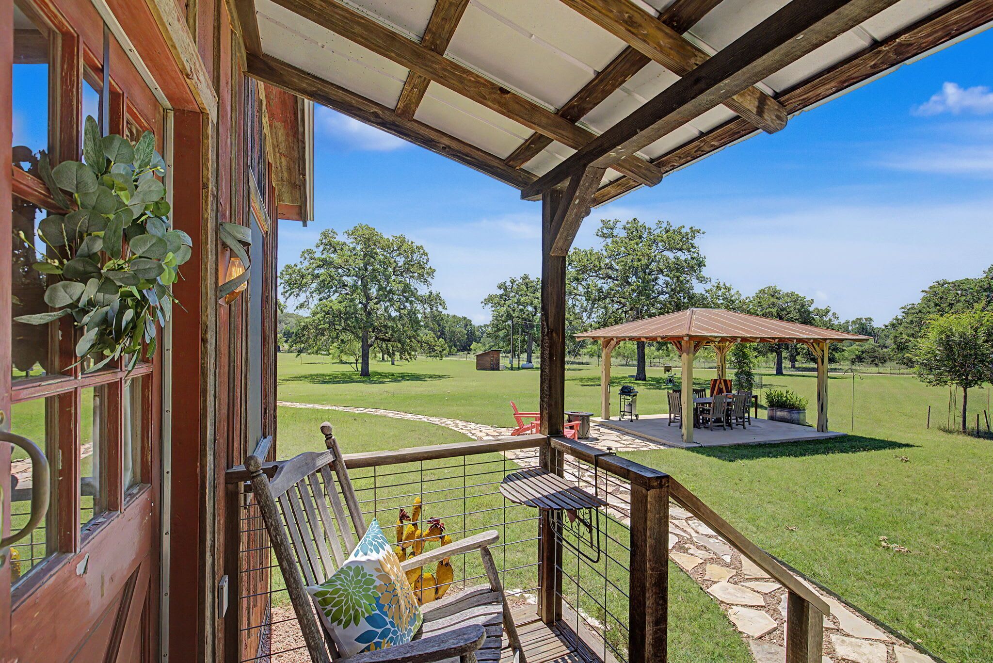 Een overdekt terras met houten vloer, een pergola en uitzicht op een grasveld met bomen.