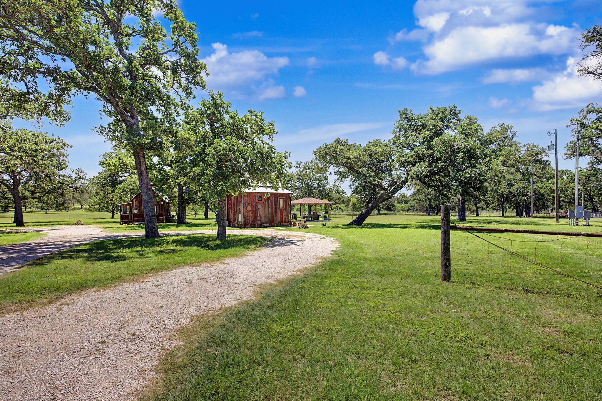 A rural setting with a gravel path leading to a rustic wooden structure, surrounded by trees and open grassy areas.