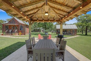 An outdoor covered patio with a wooden table and chairs, a barbecue grill, and a red barn in the background. - Cabins Out Back-nest (Fredericksburg)