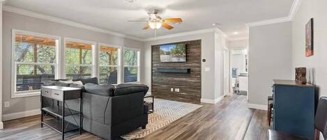 A spacious living room with a dark wood floor, a gray sofa, a ceiling fan, and a fireplace with a TV above it.