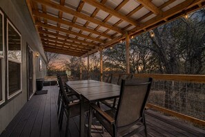 An outdoor deck with a wooden table and chairs, surrounded by a wooden railing and a view of trees. - Hye Life in the Hill Country! (Johnson City)