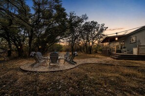 A rustic outdoor seating area with Adirondack chairs and a table, surrounded by trees and a house with a porch. - Hye Life in the Hill Country! (Johnson City)