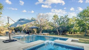 A swimming pool with lounge chairs and umbrellas, surrounded by trees. - Suniland W/pool, Hot-tub and Sauna! (Fredericksburg)