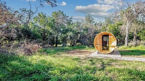 A wooden cylindrical structure with an open door, surrounded by a natural landscape with trees and grass. - Suniland W/pool, Hot-tub and Sauna! (Fredericksburg)