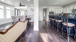 A spacious living room with a patterned sofa, a dining area with a table and chairs, a kitchen with bar stools, and a ceiling fan. - Charming Farmhouse W/hot-tub Near 290 Wine Trail (Fredericksburg)