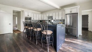 A modern kitchen with dark wood flooring, white cabinetry, a central island with bar stools, stainless steel appliances, and a tiled backsplash. - Charming Farmhouse W/hot-tub Near 290 Wine Trail (Fredericksburg)
