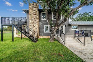 A modern two-story house with a stone chimney, a tree in the front yard, and a metal staircase leading to a balcony. - Charming Farmhouse W/hot-tub Near 290 Wine Trail (Fredericksburg)