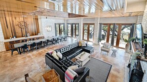 A spacious living area with a large black leather sofa, a wooden dining table, and a kitchen area with a stone wall and wooden ceiling. - Luxury Lakefront Escape on Lake Travis (Marble Falls)
