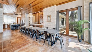 A spacious dining area with a long table and chairs, a kitchen in the background, and a view of the outdoors. - Luxury Lakefront Escape on Lake Travis (Marble Falls)