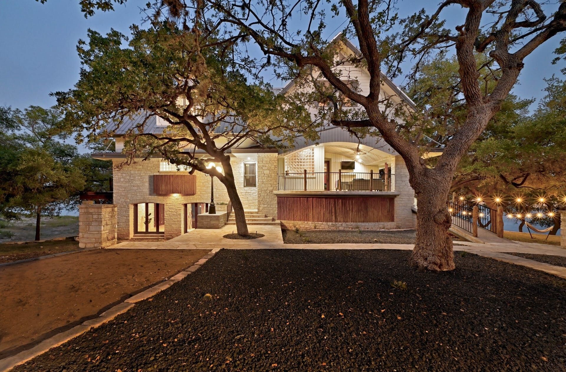 A large, two-story stone house with a wrap-around porch, a covered outdoor seating area, and a view of a body of water.