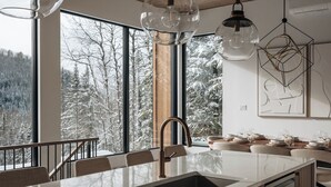 A modern kitchen with a large island, stainless steel appliances, and large windows offering a view of a snowy forest. - Chalet Symbiose21 - Spa& Sauna& Ski (Saint-Come)