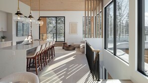 A modern kitchen with a central island, bar seating, and large windows offering a view of a snowy landscape. - Chalet Symbiose21 - Spa& Sauna& Ski (Saint-Come)