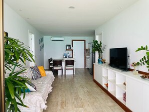 Ein Wohnzimmer mit einer Couch, einem Fernseher und Regalen. Im Hintergrund befindet sich ein Essbereich mit Stühlen und einem Tisch.