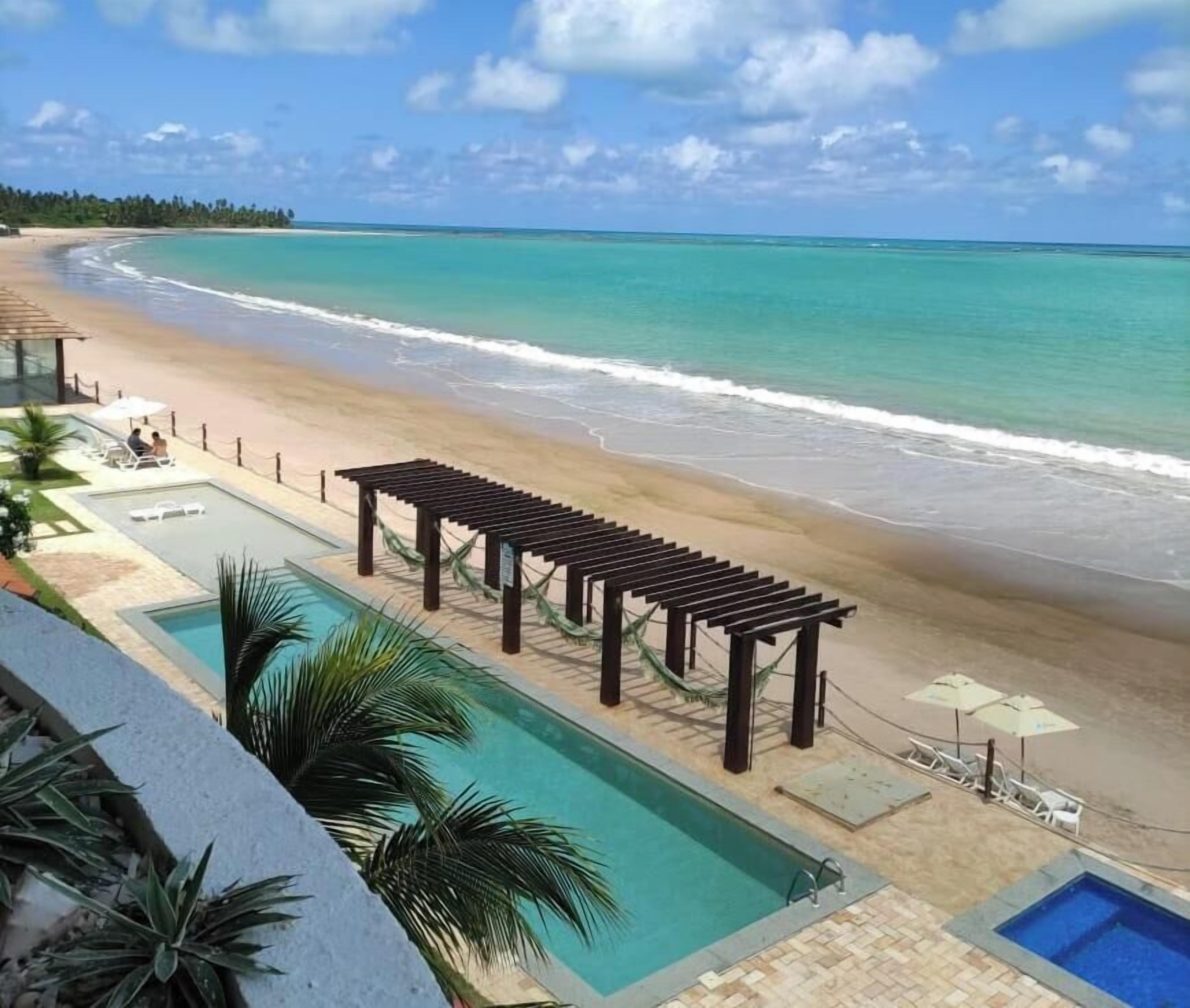 Strandudsigt med pool, et træbeklædt pergola og en klar himmel.