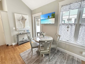 A dining area with a round table, chairs, a rug, a TV, and a window with curtains. - Seaglass Village 217 - Barefoot Escape: Cozy 2-bedroom condo in Wells, Maine. (Wells)