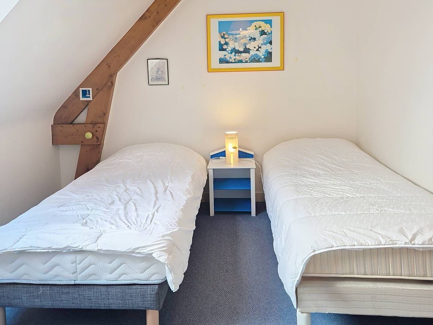 Une chambre avec deux lits simples, une table de chevet avec une lampe, un tableau encadré au mur et une structure en poutres apparentes.