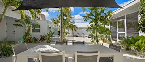 Ein überdachter Essbereich im Freien mit Tisch und Stühlen, umgeben von tropischen Pflanzen und einem Pool im Hintergrund.