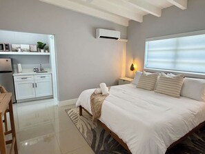 A bedroom with a large bed, a bedside table, a nightstand, a window with blinds, a wall-mounted air conditioner, and a kitchenette with a microwave, sink, and cabinets. - 3 Bedroom on the Barrier Island. Walk to the Beach (Fort Lauderdale)