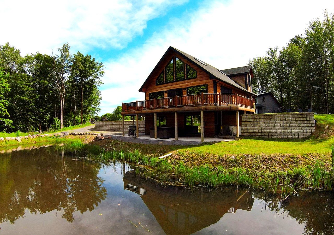 A wooden cabin with a balcony, a pond in front, and a stone wall.