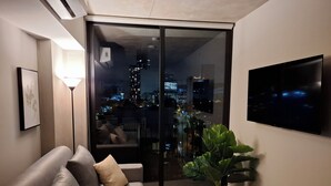 Una sala de estar moderna con un sofá, una planta grande en maceta, un televisor de pantalla plana y un ventanal corredero que da a un balcón con vistas a la ciudad.