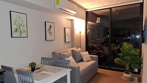Una sala de estar moderna con un sofá gris, una mesa de comedor blanca y sillas. Hay grabados botánicos enmarcados en la pared, un amplio ventanal con vistas a la ciudad y una planta en maceta en el suelo.