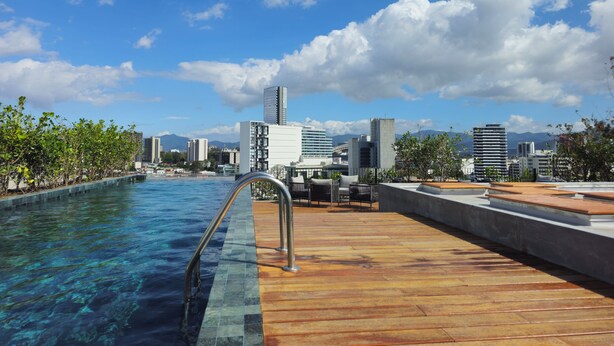 Una piscina en la azotea con agua cristalina, terraza de madera y un horizonte urbano al fondo.