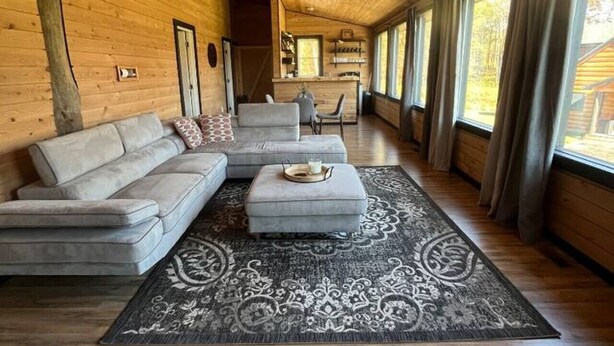 A spacious living room with a wooden ceiling, a grey sectional sofa, a matching ottoman, and a patterned rug. - Meadow Ridge Apartment — Minutes to Salmon River (Altmar)