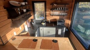 A compact kitchen with wooden walls, a central island with two sinks, and a window providing a view of the outdoors. - Meadow Ridge Apartment — Minutes to Salmon River (Altmar)