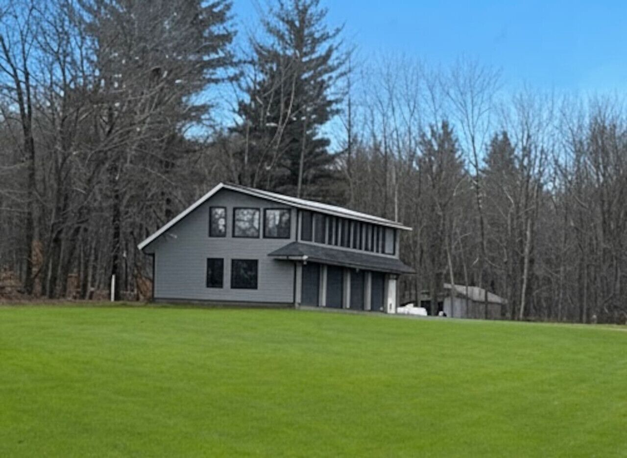 A modern two-story building with a flat roof and large windows, surrounded by a grassy field and trees.