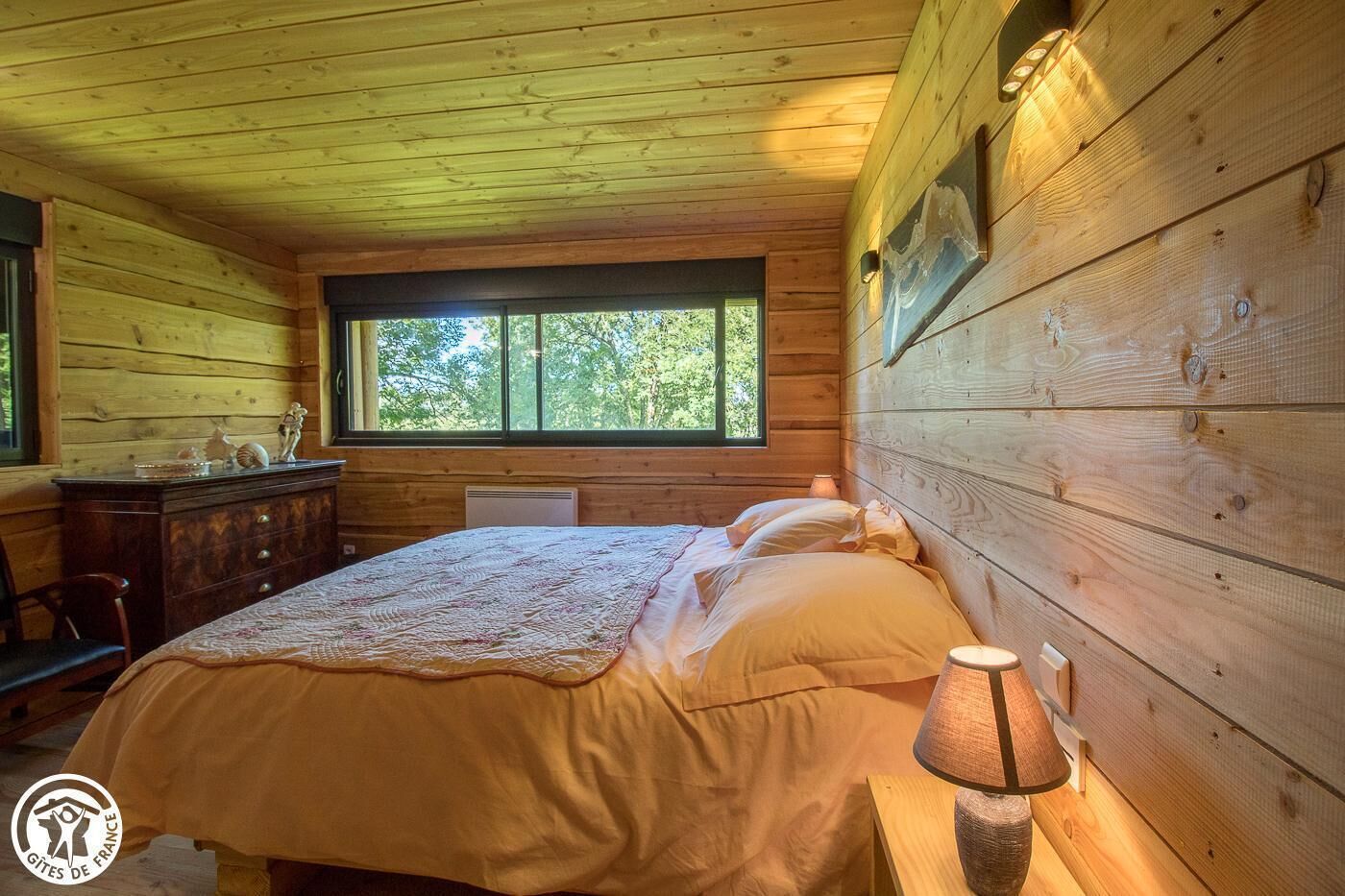 A wooden cabin bedroom with a bed, dresser, and a window with a view of trees.