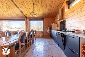 Ein Holzblock-Innenraum mit Esstisch, Stühlen, einer Küche mit modernen Geräten und großen Fenstern.