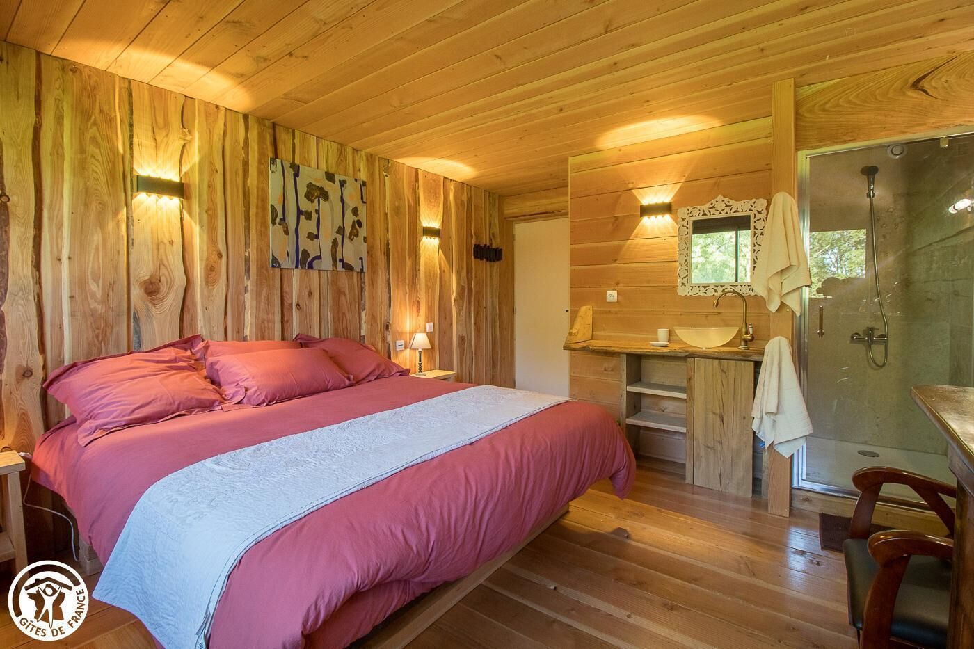 Ein Schlafzimmer in einer Holzhütte mit Bett, Nachttischlampen, einer Holzkommode und einem Badezimmer mit Waschbecken und Dusche.