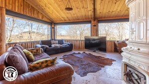 L’intérieur d’un chalet rustique est agrémenté d’un canapé en cuir, d’un téléviseur, d’une cheminée et d’une grande fenêtre donnant sur un paysage naturel.